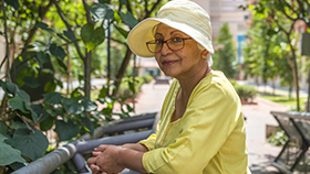 An elderly woman leaning on a fence in a garden setting.